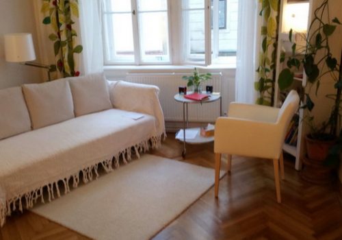 Ein Wohnzimmer mit einer weißen Couch und einem Fenster bietet eine ruhige Atmosphäre für Craniosacral-Therapie oder somatische Traumatherapie.